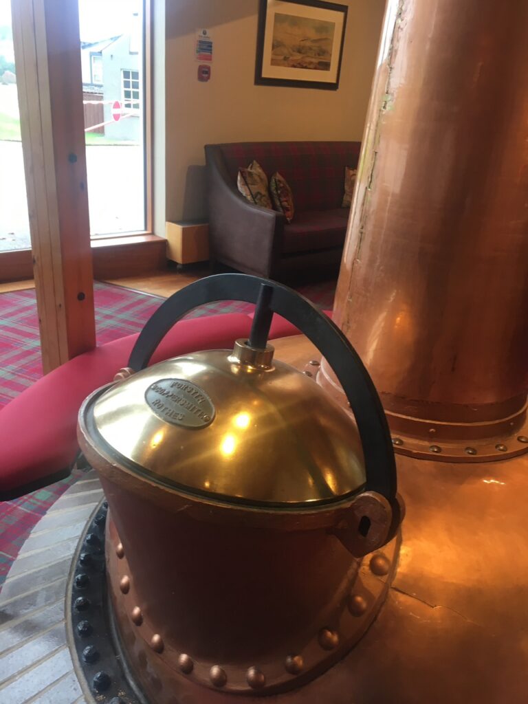 グレンファークラス蒸留所のロビーに置かれた、ポットスチルを模した暖炉のような美しいオブジェ。背景の大きなガラス窓からは、パノラマに広がる緑豊かなお庭が見える