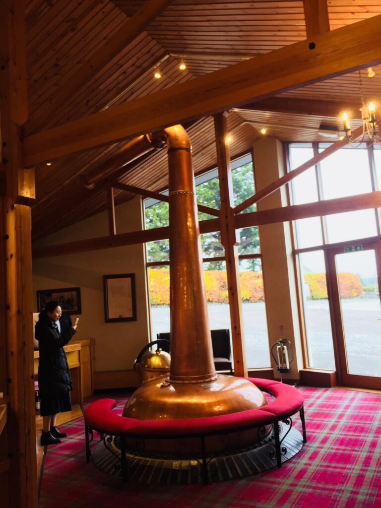 グレンファークラス蒸留所のロビーに置かれた、ポットスチルを模した暖炉のような美しいオブジェ。背景の大きなガラス窓からは、パノラマに広がる緑豊かなお庭が見える