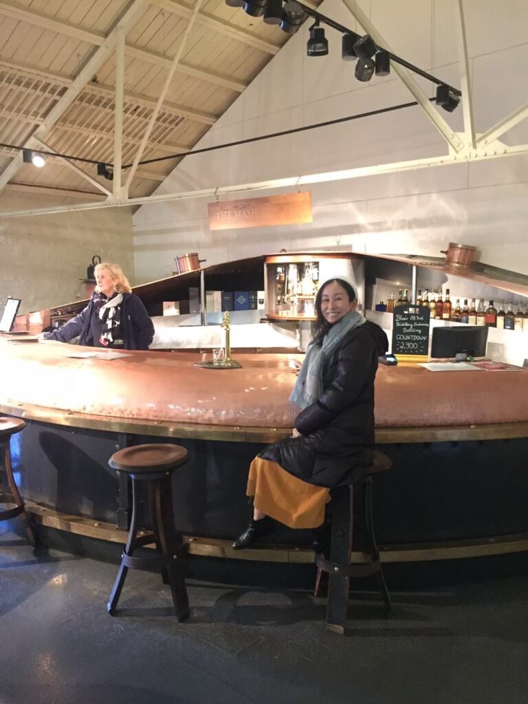 ブレアソール蒸留所のビジターセンター内「マッシュ・タン・バー」に座る筆者。クライヌリッシュ蒸留所で実際に使われていた巨大な銅製・鋳鉄製の糖化槽をアップサイクルした、世界でも類を見ないテイスティングバー。足元の濾過プレートや攪拌機をイメージした内装など、ウイスキー製造設備を再利用した革新的な設計。スコットランド・ピトロッホリーの蒸留所体験。