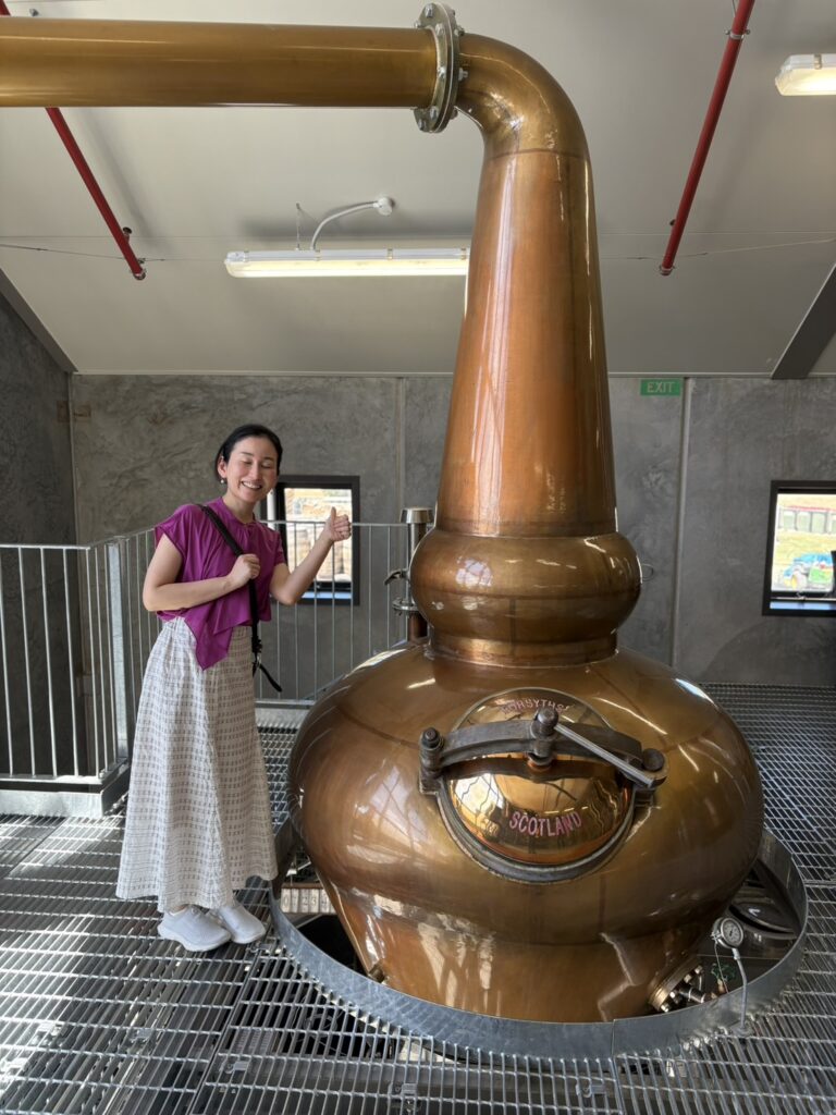 伝統的な形状のポットスチルを背に、カードローナの職人魂に触れて笑顔を見せる筆者（バー店主）。銅の輝きが蒸留所の歴史と情熱を物語る一枚