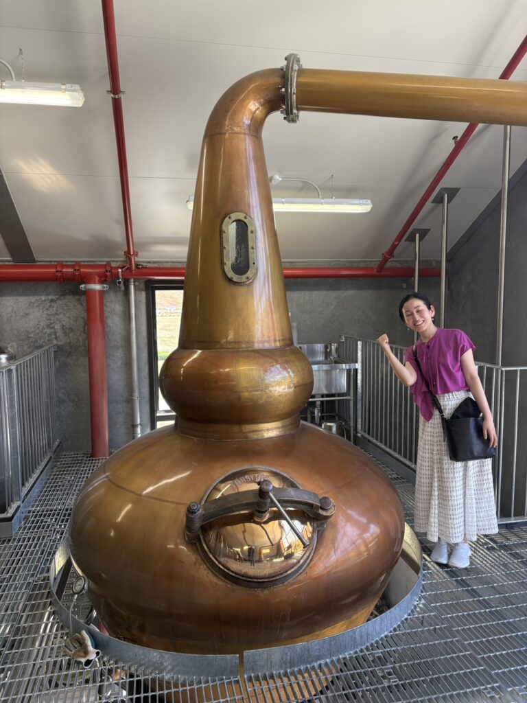 伝統的な形状のポットスチルを背に、カードローナの職人魂に触れて笑顔を見せる筆者（バー店主）。銅の輝きが蒸留所の歴史と情熱を物語る一枚