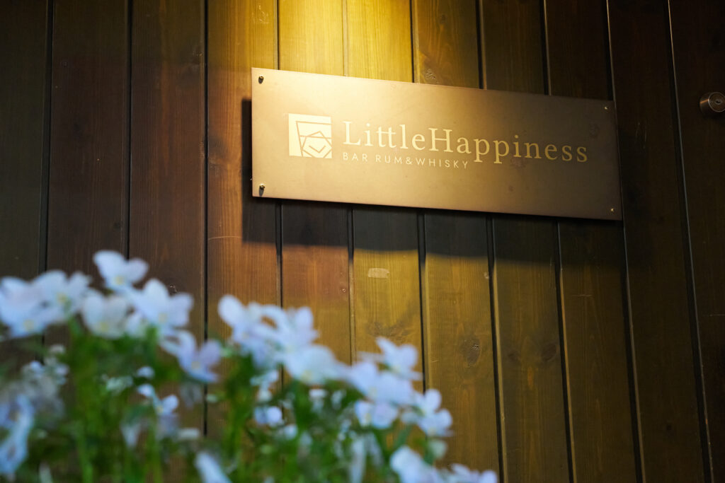 The exterior sign of Bar Little Happiness in Nagarekawa, Hiroshima, surrounded by flowers